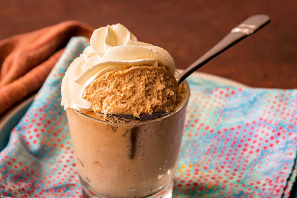 A glass of butterscotch mousse topped with whipped cream with a spoon in it.