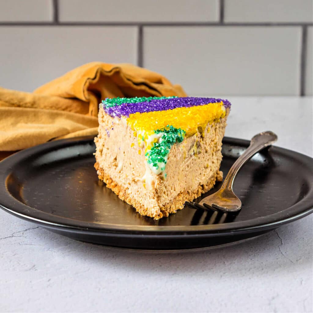 A slice of King cake cheesecake on a black plate with a fork.