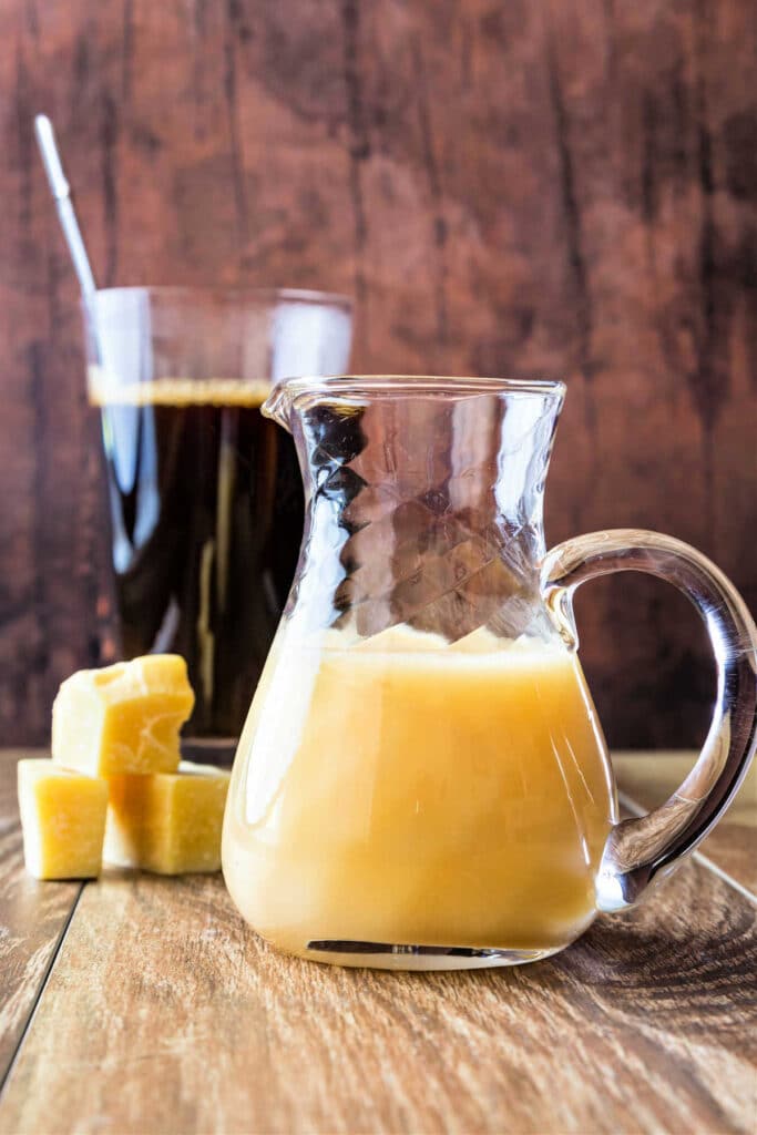 A small glass pitcher of white chocolate sauce with chunks of pure cocoa butter to the side and a glass of coffee in the background.