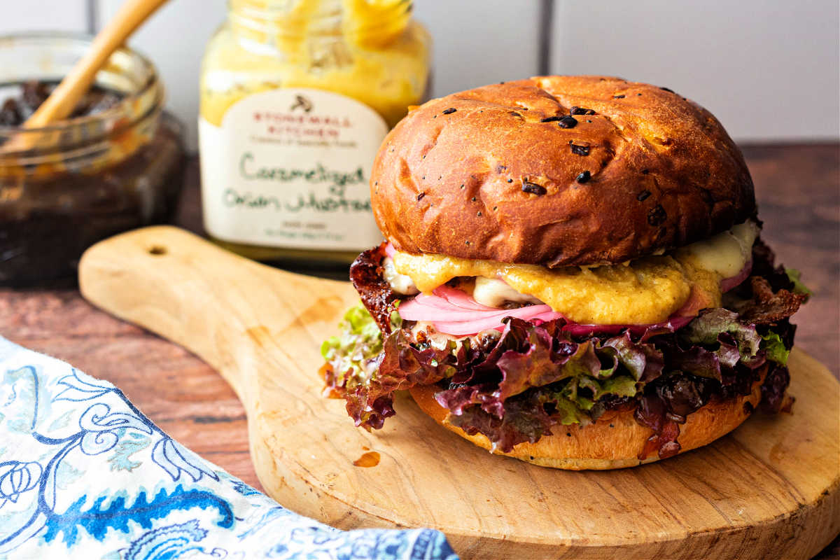 A burger on a wooden board with a jar of mustard and one of onion jam.