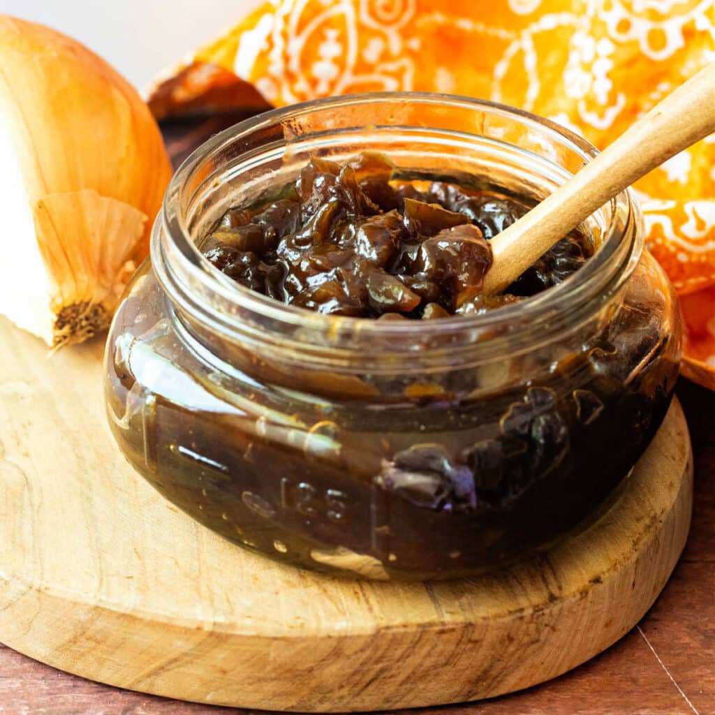 A jar of onion jam on a wooden cutting board with a wooden spoon
