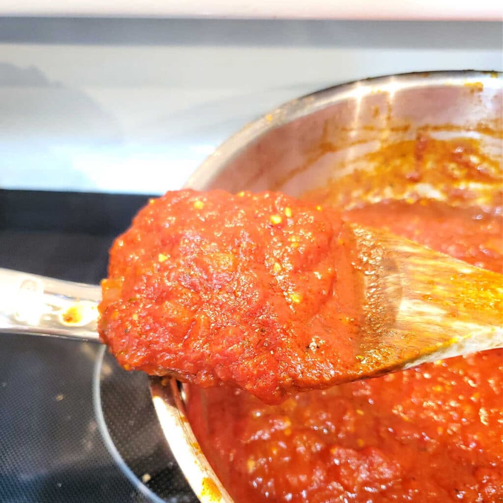 A close up of a metal pan of pizza sauce with some of the sauce mounded on a wooden spoon balanced on the rim of the pan.