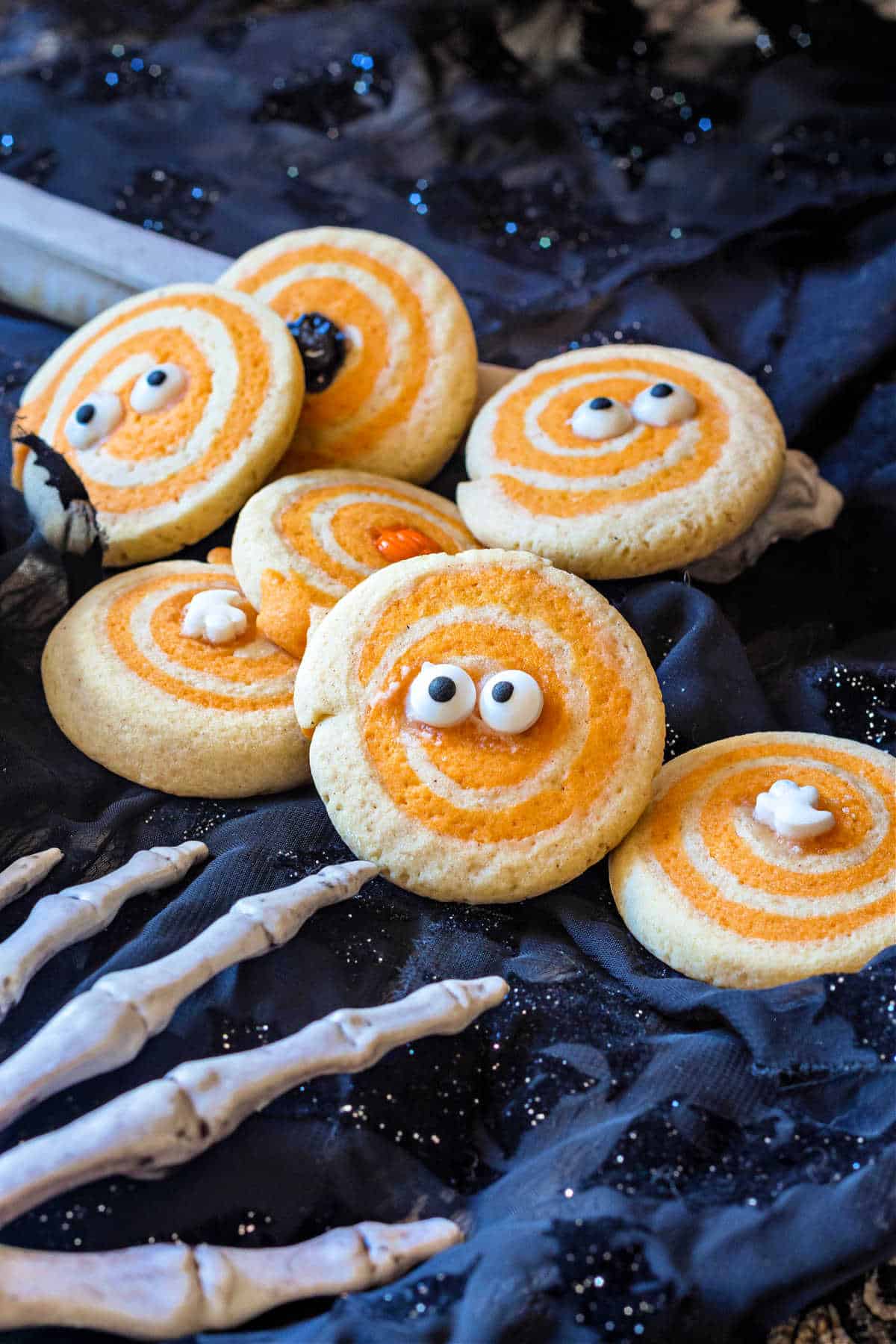 Halloween sable cookies, each with a different Halloween decoration in the center, shot on a black backgroun with a gray plastic, skeletal hand in the foregound.