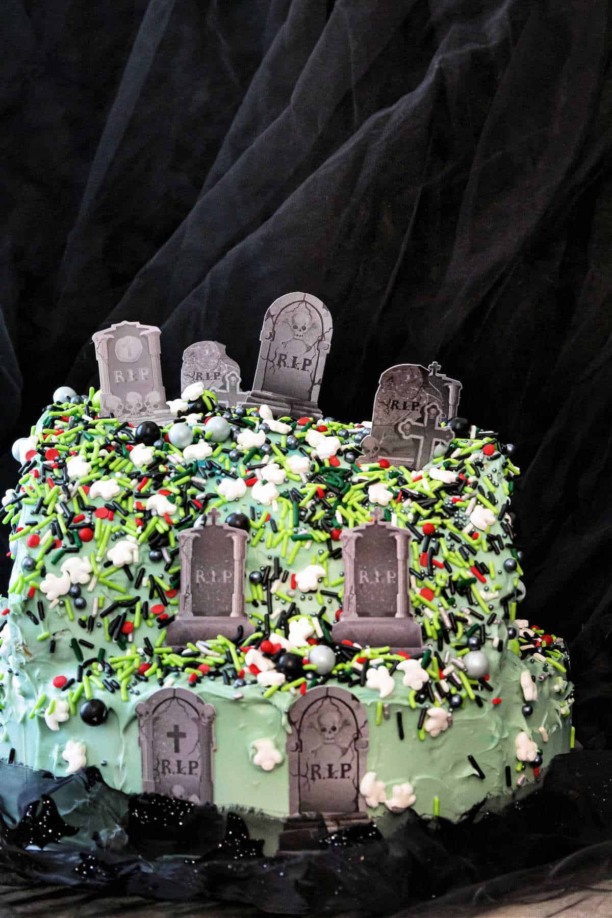 A "hilltop graveyard cake" that is iced in blue-ish gray frosting with multicolored sprinkles. It has small, gray, cardstock headstones stuck in it at intervals so it looks like a graveyard.