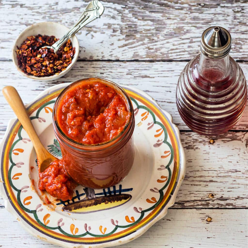 A square image of a jar of pizza sauce, some vinegar in a glass bottle, and a small dish of pepper flake.