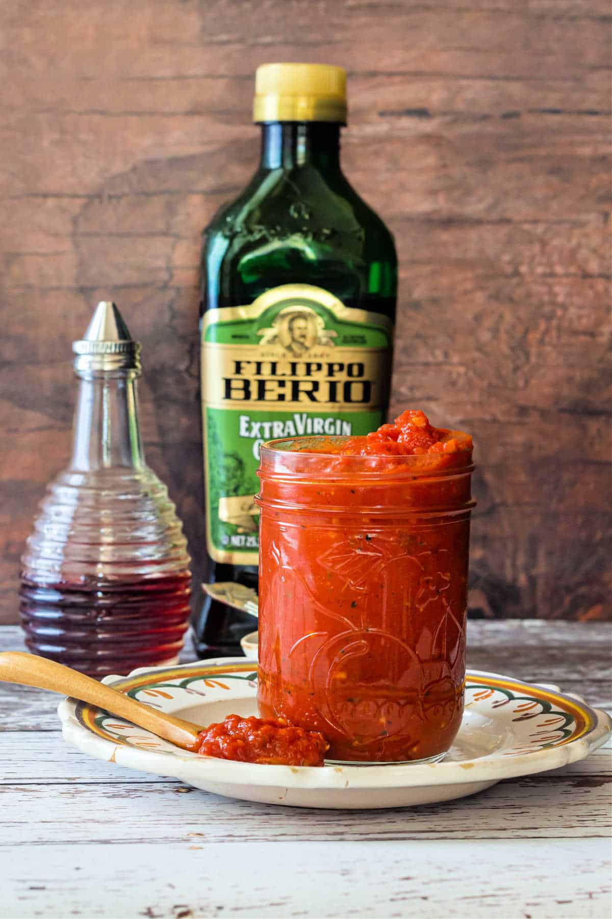A glass jar of pizza sauce on a plate with a cruet of red wine vinegar and a large green bottle of olive oil behind it.