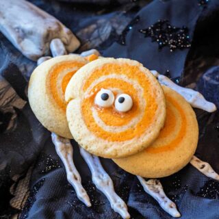 Three orange and white swirled cookies, one with googly eyes, sitting on a plastic skeletal hand on a black background.