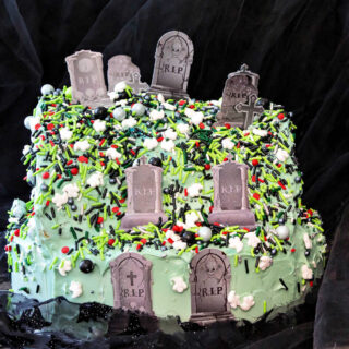 A cake decorated with multi-colored sprinkles in the shape of a half circle so it looks like a hill. There are paper headstones stuck in it at intervals so it looks like a graveyard on a hill.
