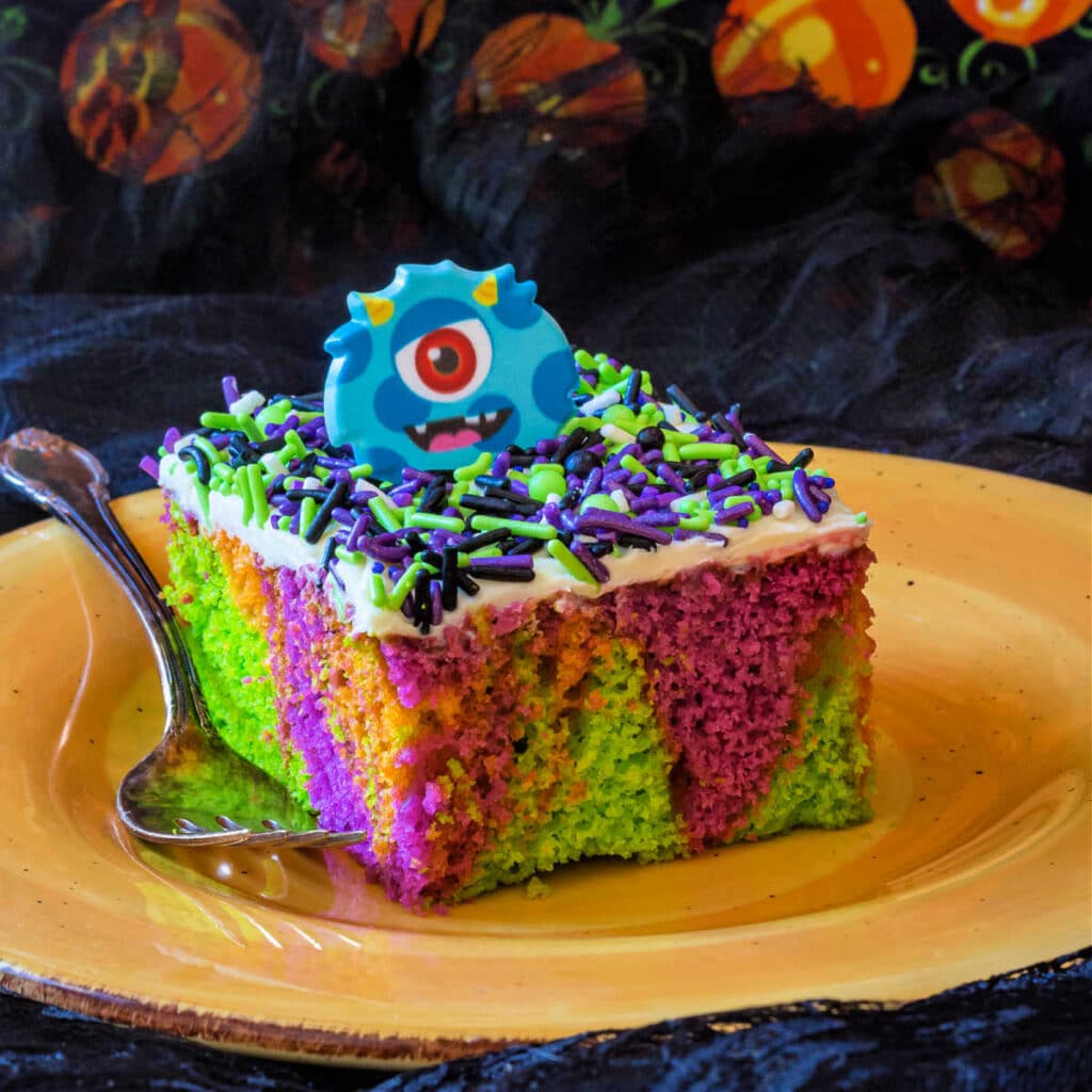 A slice of monster cake. The cake is swirled purple, orange, and green, and the top is ices with white frosting covered with lots of black, green, and purple sprinkles.