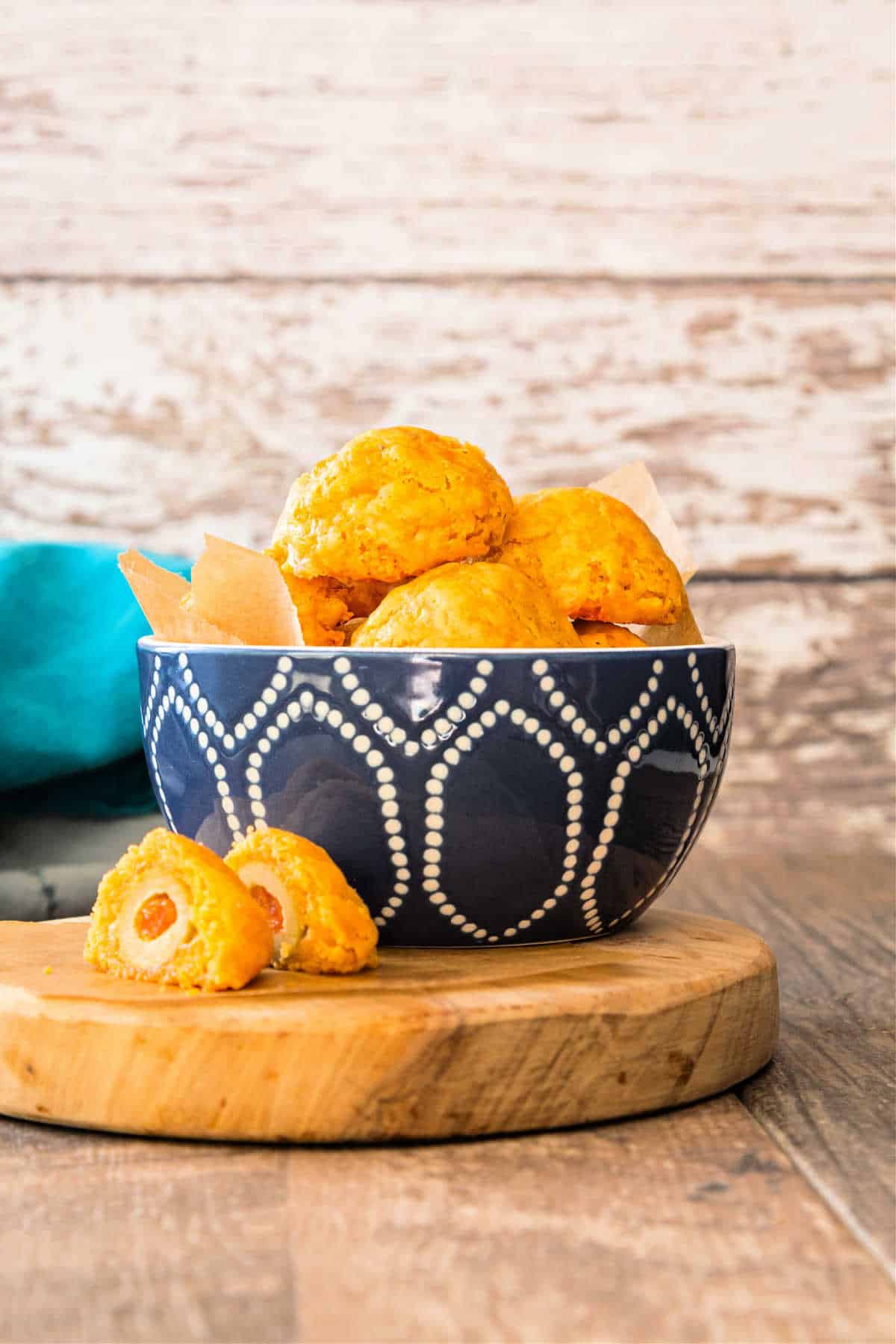A blue pottery bowl filled with orange cheese puffs. The bowl is on a cutting board with a cheese puff on it cut in half to reveal a pimento-stuffed green olive inside.