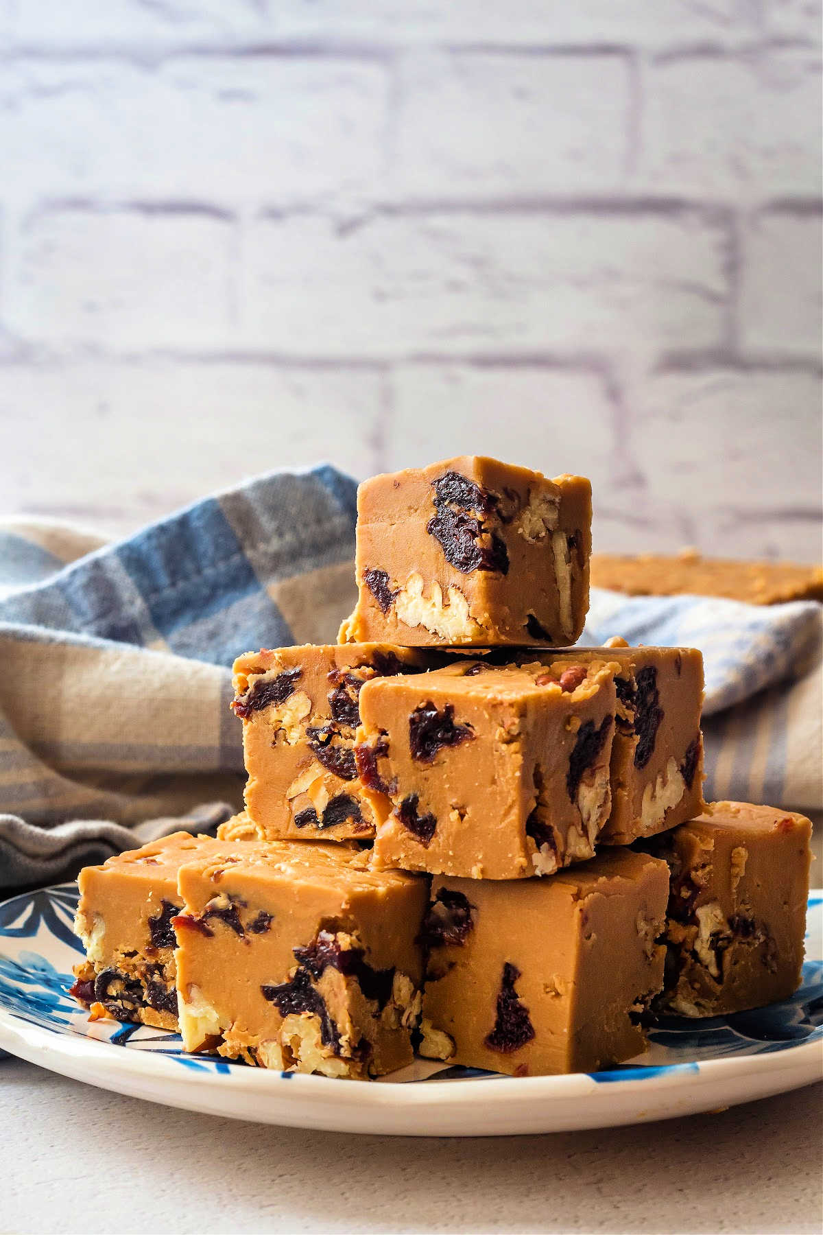 A blue plate with a stack of penuche fudge candies on it. The brown sugar fudge candies are studded with dried cherries and toasted pecans.