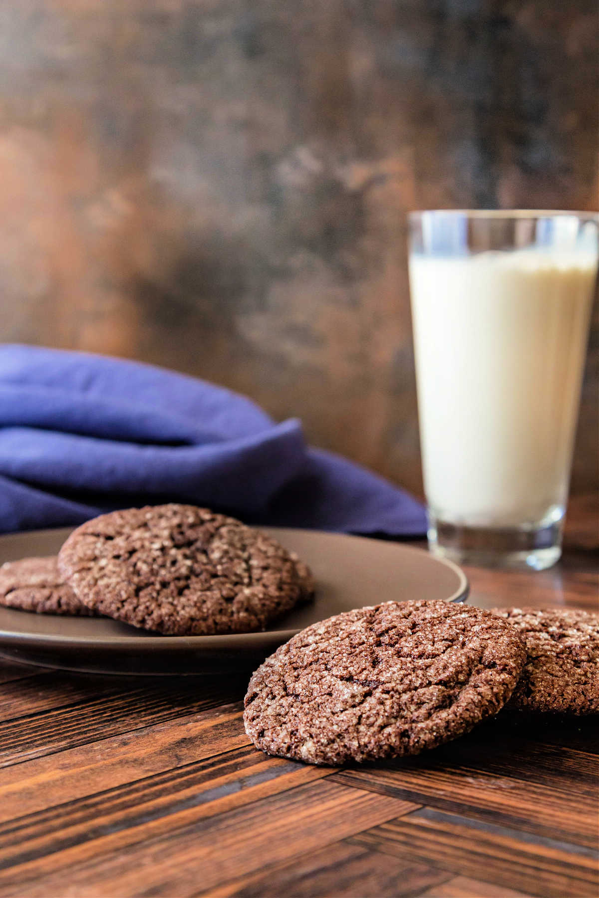 A glass of milk with chocolate cookies and a blue napkin shot on a wooden surface with a brown and blue mottled backdrop behind.