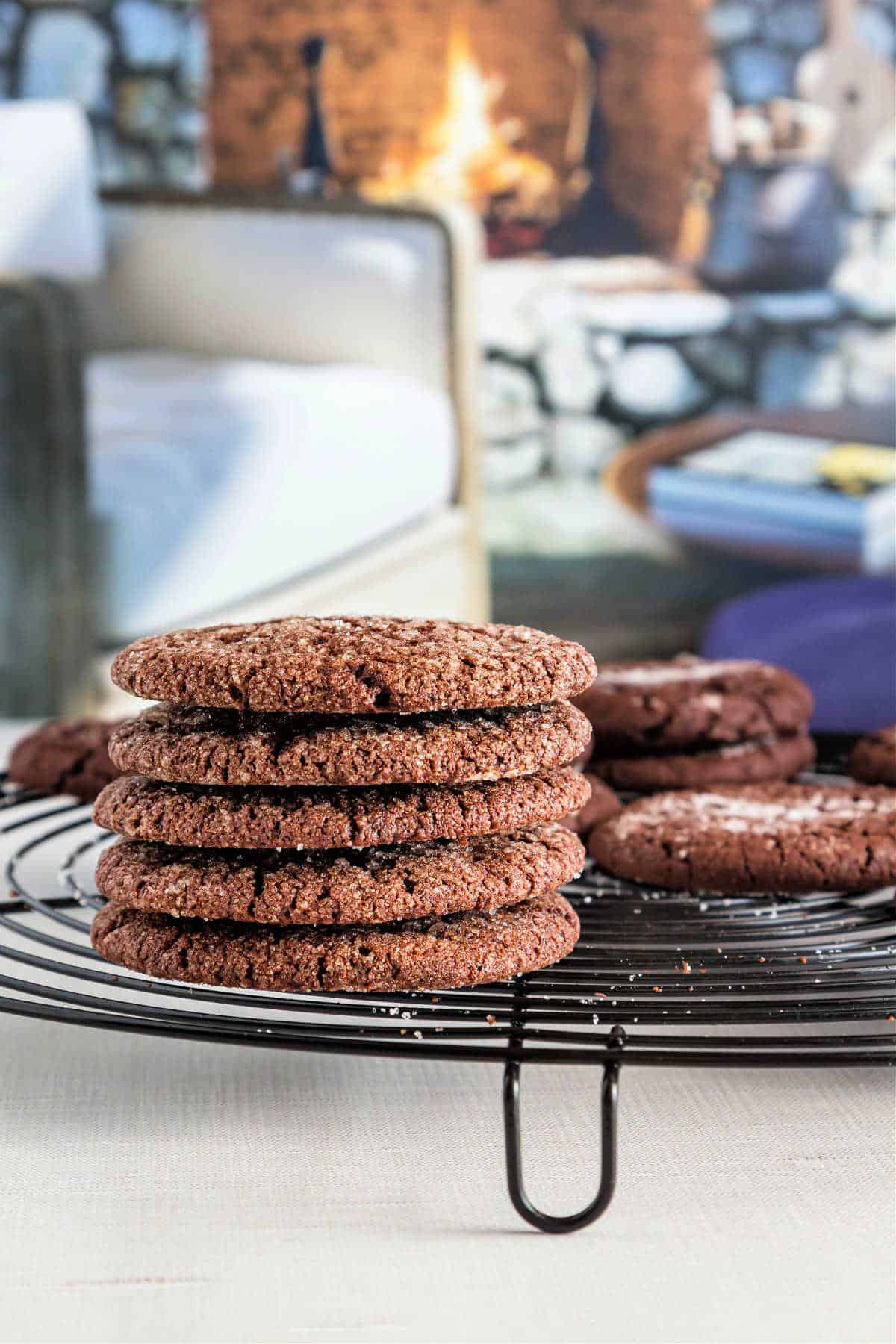 A stack of crisp cocoa cookies on a black cooling rack with a fireplace and chair in the background.