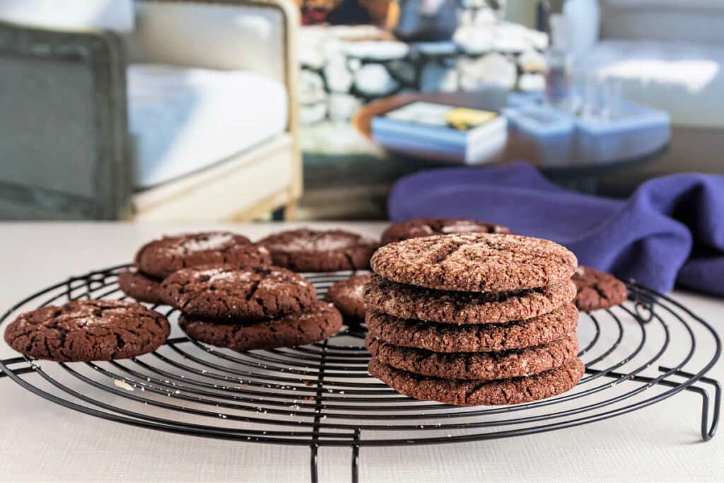 A round, black wire cooling rack with cocoa cookies cooling on it on a cream-colored surface with living room furniture in the background.