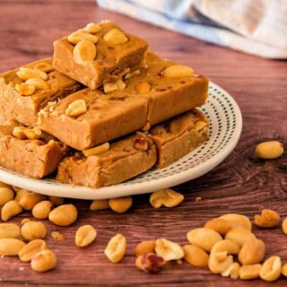 A small plate with a pile of homemade honey peanut butter fudge with peanuts scattered around it shot on a walnut wood-colored surface.