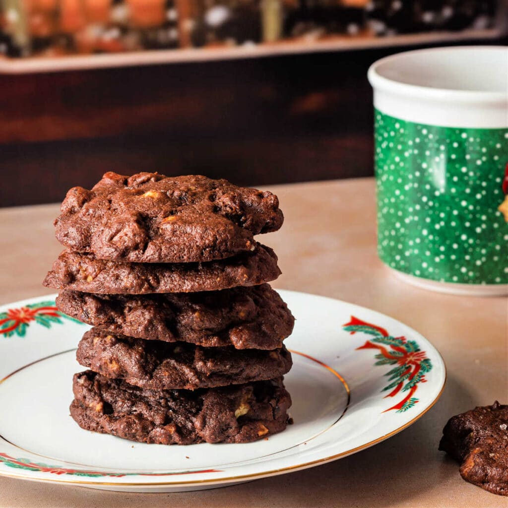 A white plate with poinsettias on it with a stack of cookies and a green mug in the background.
