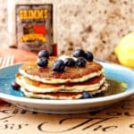 A close shot of stack of three pancakes on a blue plate topped with a pat of butter and fresh blueberries.