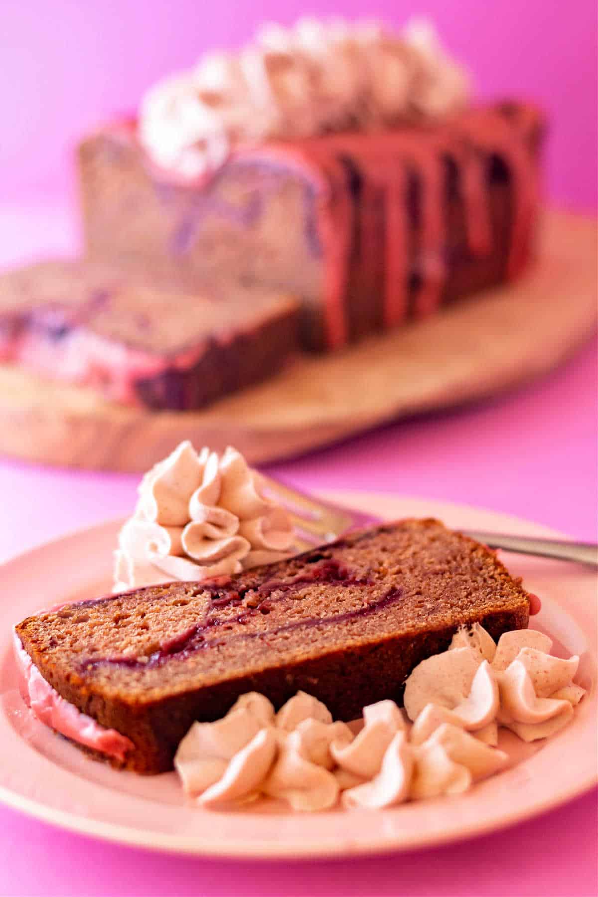 A slice of strawberry cake with a strawberry swirl with strawberry whipped cream on a pink plate with the rest of the cake in the background.