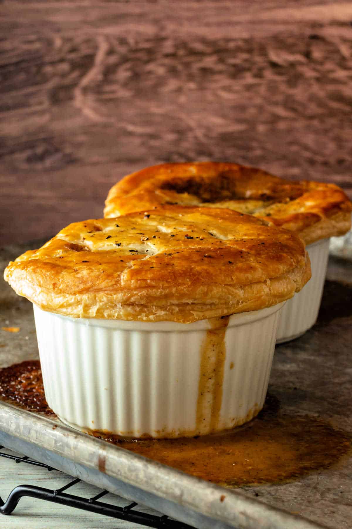 White, puff pastry-topped ramekins cooling on a rack. There is a little bit of gravy running down the side of the ramekin in the front.