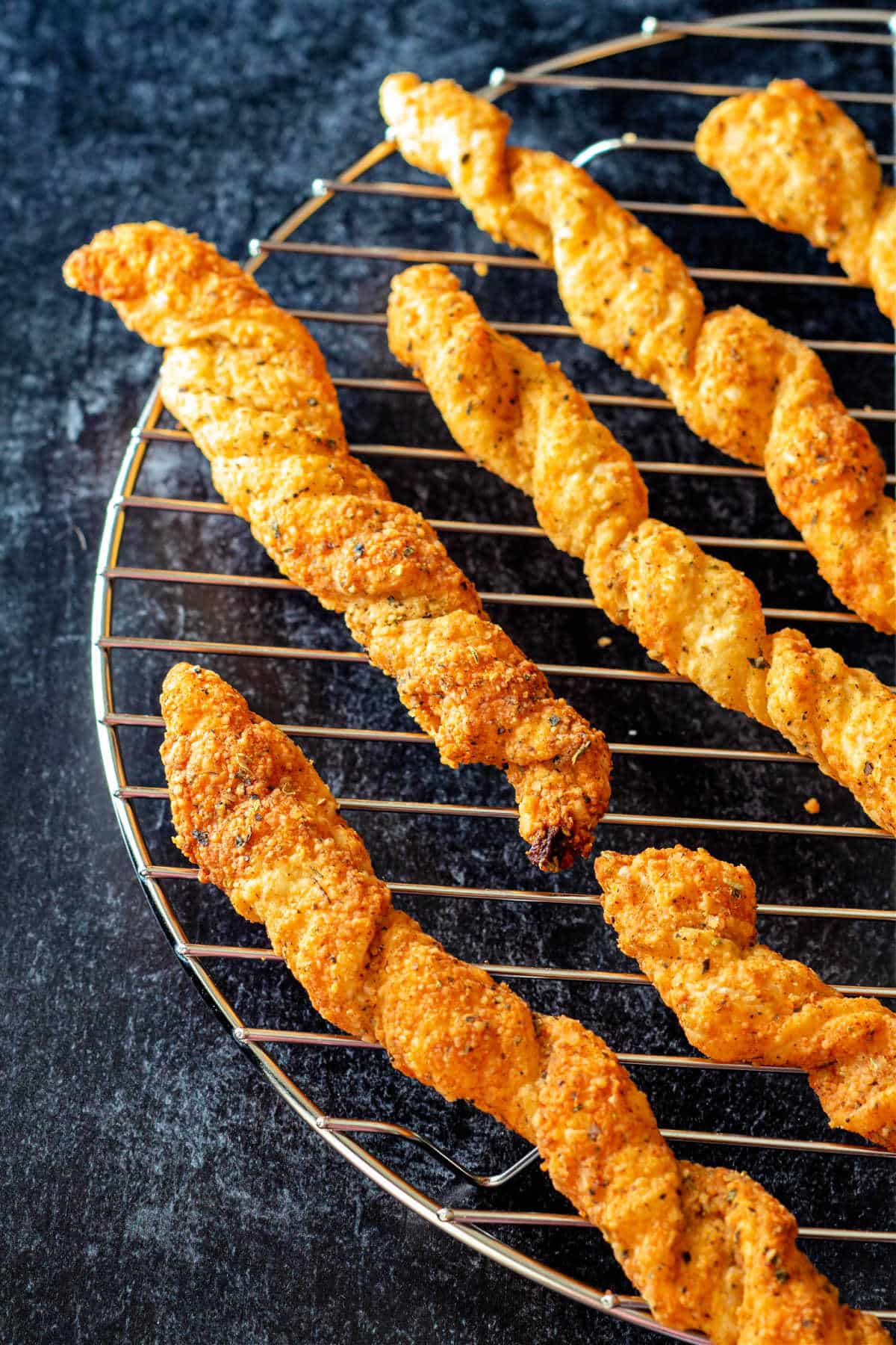 Puff pastry cheese straws cooling on a rack on a black surface.