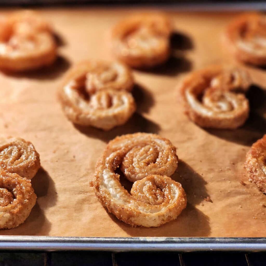 A close up shot of palmiers in the oven. They are puffed out to resemble ears, and there is coarse sugar embedded in the dough. They are golden brown and a little shiny.