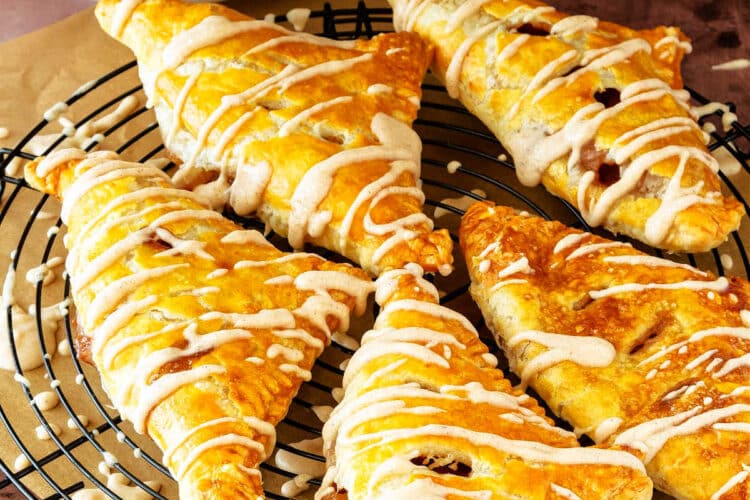 A black cooling rack with five puff pastry apple turnovers on it. They have just been glazed, and glaze is dripping off onto parchment below the rack. The white bowl of glaze with a whisk in it is in the background.