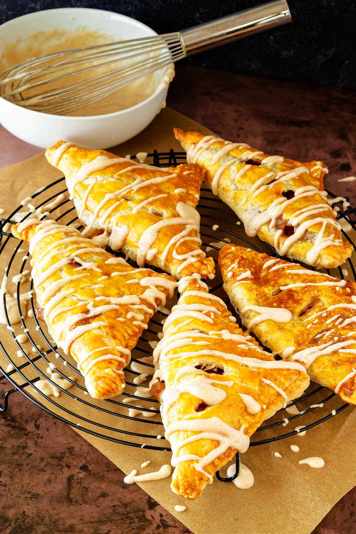 A black cooling rack with five puff pastry apple turnovers on it. They have just been glazed, and glaze is dripping off onto parchment below the rack. The white bowl of glaze with a whisk in it is in the background.