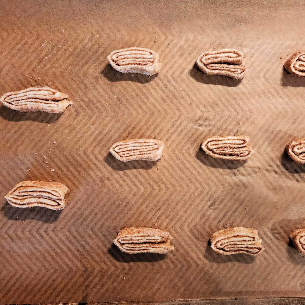 An overhead shot of several folded palmiers on parchment before baking. They are thin, and there are lines of darker sugar between the folds of the lighter pastry. They are not at all puffed before baking.