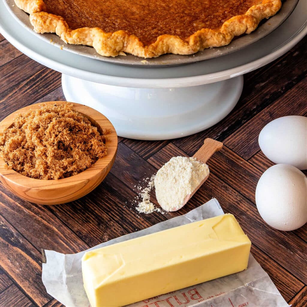 A stick of butter, a bowl of dark brown sugar, a small scoop of cornmeal and two white eggs on a wooden surface. You can see an edge of a baked pie on a white pedestal at the top of the frame as well.