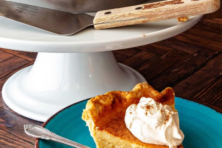 A chess pie in a pie pan on a white pedestal with a slice of pie on a blue plate with whipped cream on top.