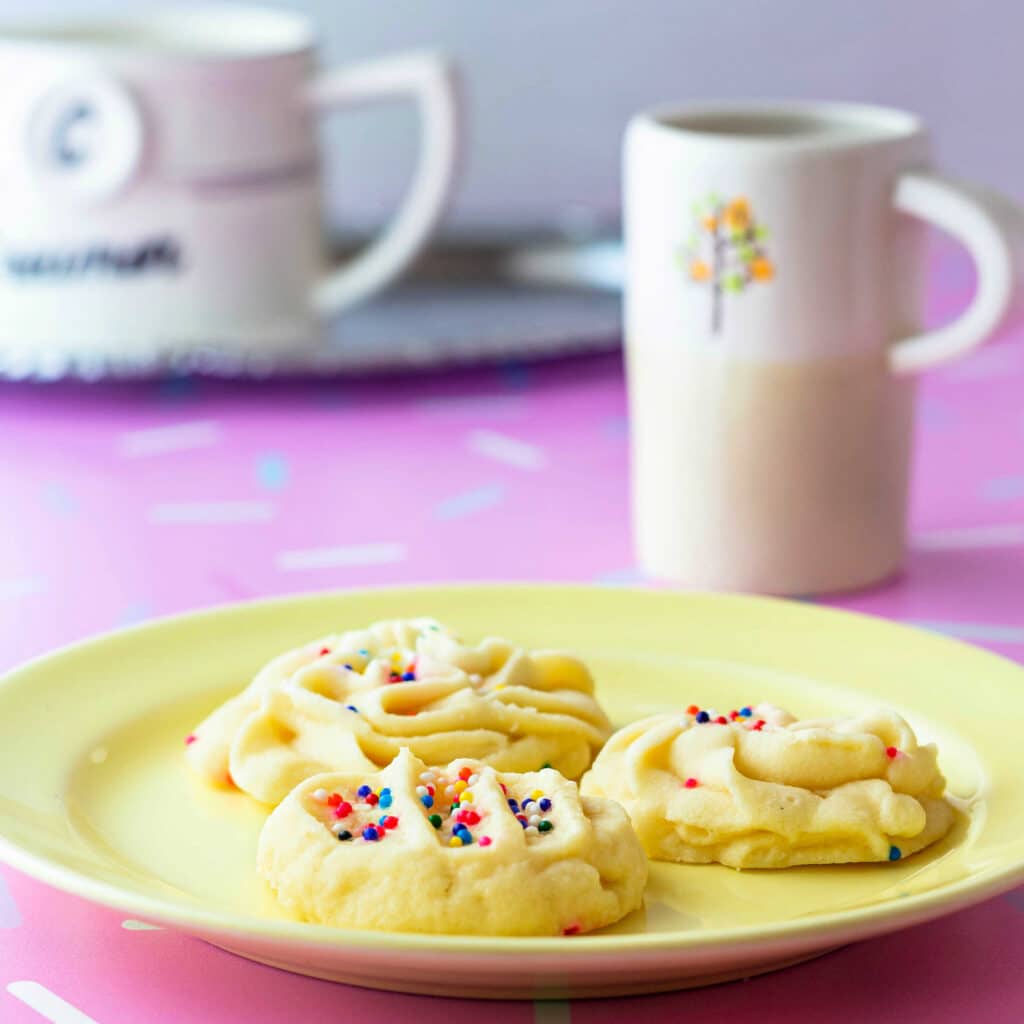 A yellow plate with 3 cookies on it with sprinkles, a small espresso cup, and a creamer in the background.