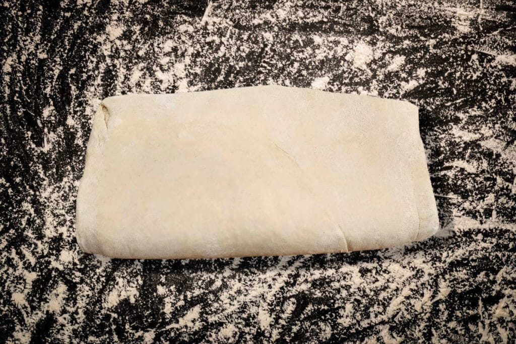 An overhead shot of folded puff pastry on a flour-dusted black surface.