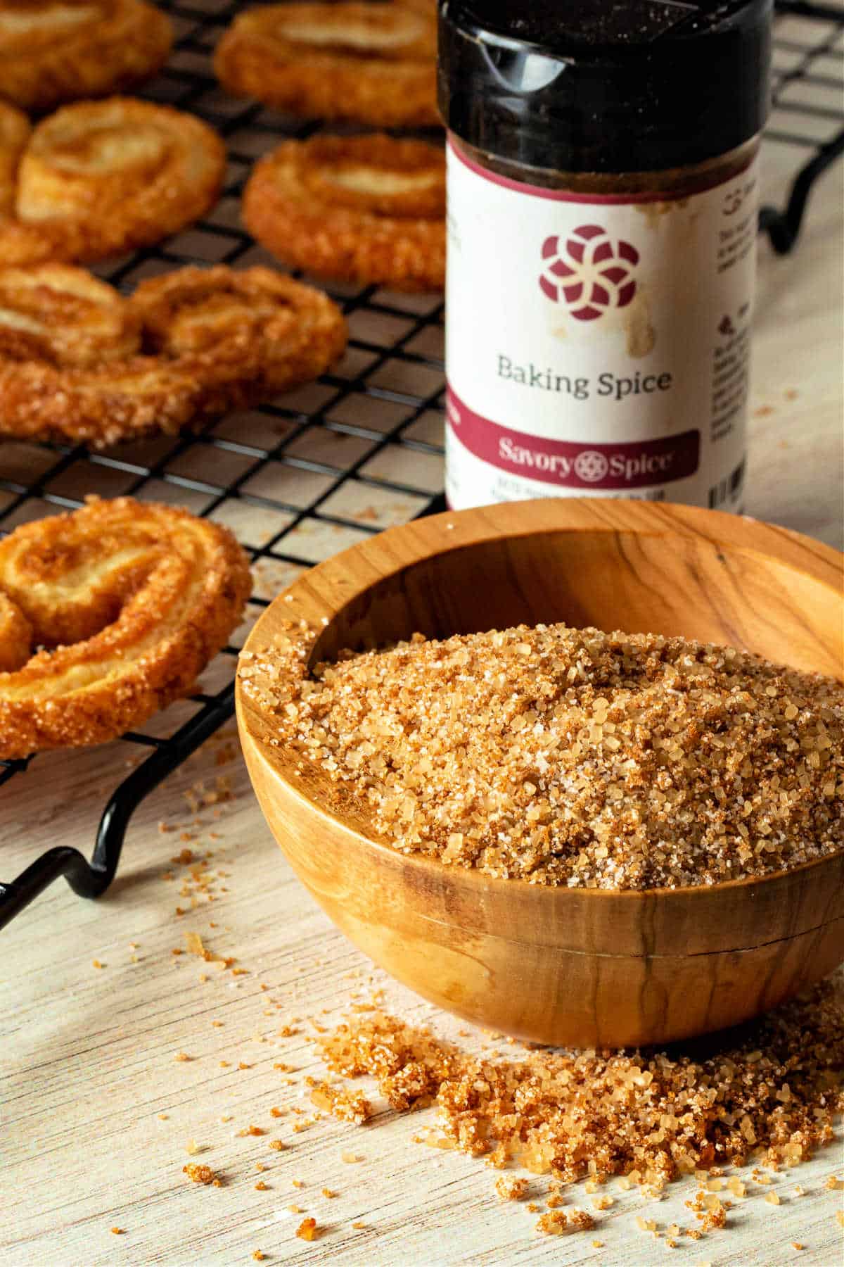 A wooden bowl of mixed sugar and spice with a jar of baking spice behind it and a metal cooling rack of palmiers.