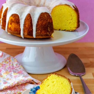 A lemon Bundt cake on a white cake stand with a slice of the cake on a plate.