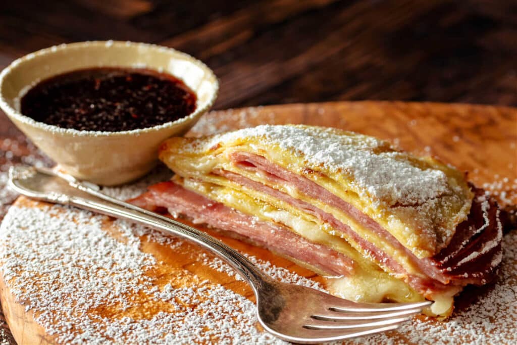 A baked Monte Cristo crepe dusted with powdered sugar with a small dish of raspberry jam next to it.