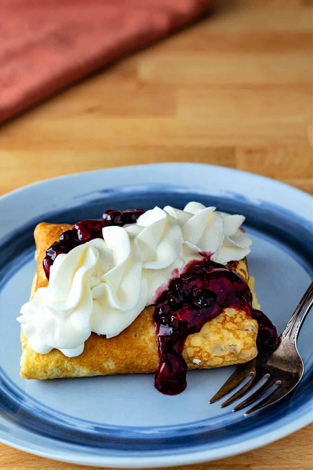 A crepe folded into a pillow shape topped with berry sauce and whipped cream. The crepe is on a blue plate with a fork to the right.
