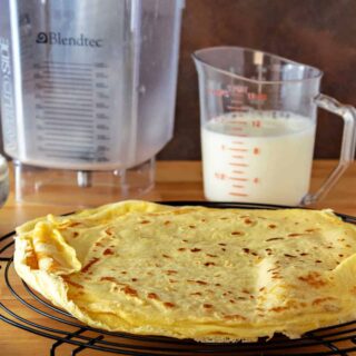 A round cooling rack with a stack of crepes on it. There's a clear plastic pitcher half-filled with milk and an empty blender jar behind the crepes.