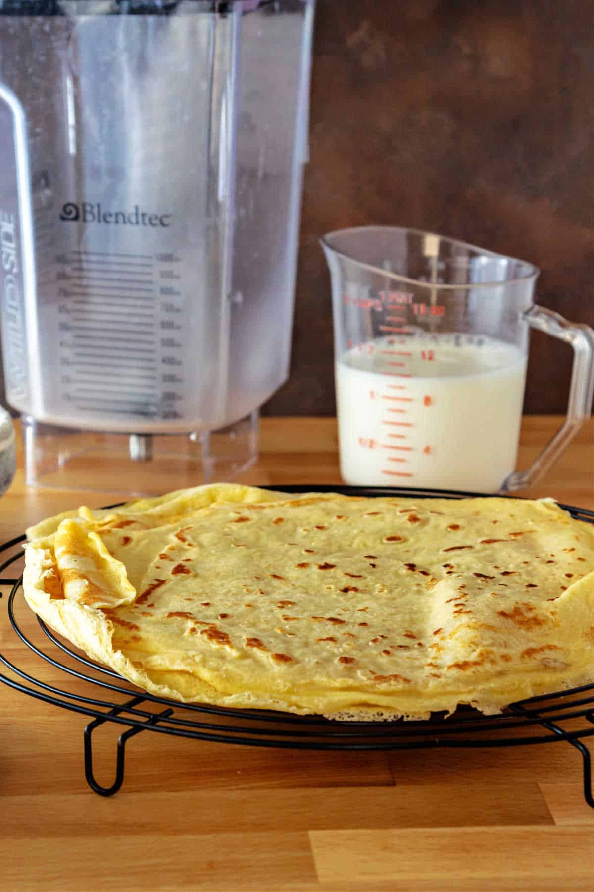 A round cooling rack with a stack of crepes on it. There's a clear plastic pitcher half-filled with milk and an empty blender jar behind the crepes.