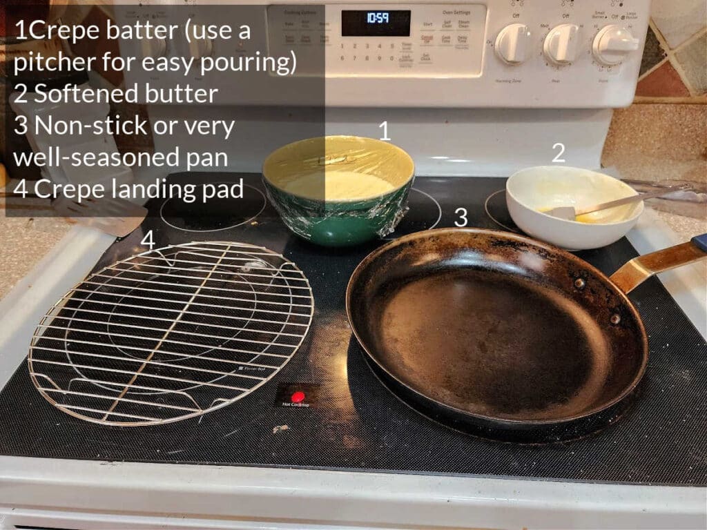 The top of a black glass stove with a nonstick pan on it, a cooling rack, a green bowl of crepe batter, and a white bowl of butter. Text overlay labels each one. This is a crepe-making station set-up.