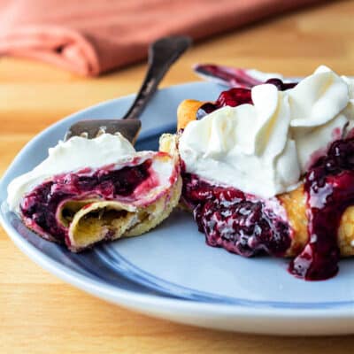 A blue plate with a crepe with mixed berry and whipped cream filling. The crepe is cut open and a bite in on a fork.