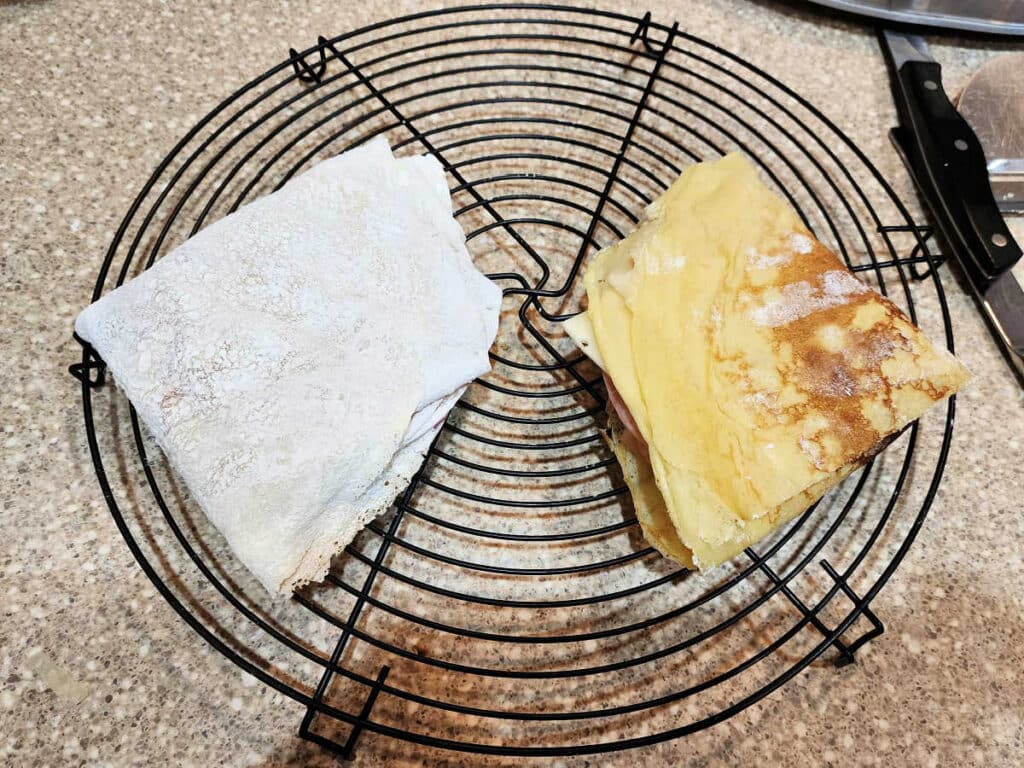Two crepes folded into triangles on a round cooling rack. One has been dusted with cornstarch.