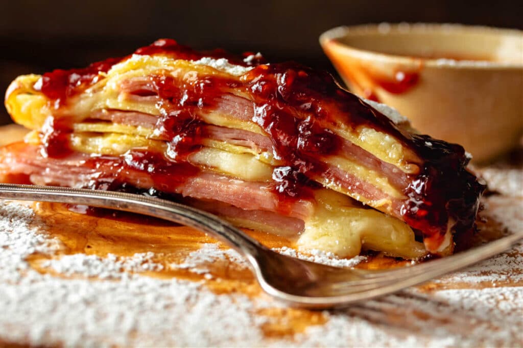 A close up of a folded crepe cut open to show layers of crepe, melted cheese, ham and turkey with some raspberry jam drizzled over the top.