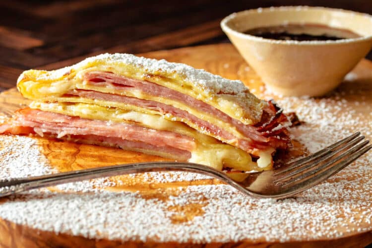 A baked Monte Cristo crepe with the layers of ham, turkey, and melted cheese showing. There is powdered sugar dusted over the crepe with a small bowl of raspberry jam and balsmic vinegar in the background.