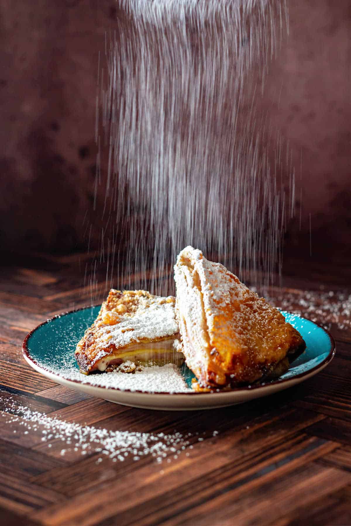 A vertical image of powdered sugar showering down over the top of a deep fried crepe filled with ham, turkey, and Swiss cheese. The crepe is cut in two and one half is leaned against the other half. Both sit on a blue plate with a brown rim.
