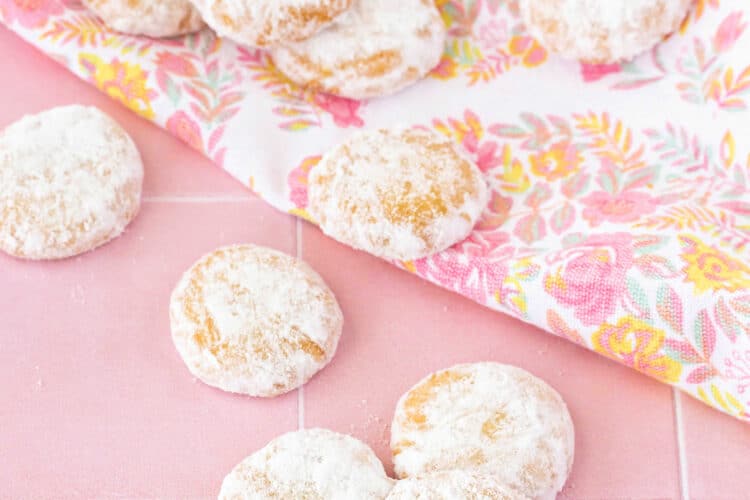 Small round cookies covered with powdered sugar on a peach tile background with a floral kitchen towel.