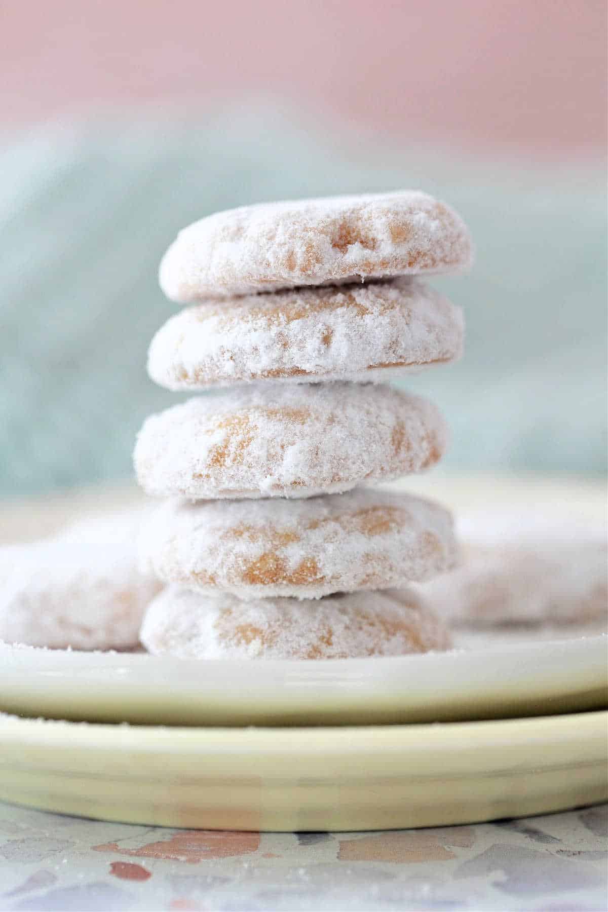 A stack of five small round cookies dusted with powdered sugar and shot on a pale yellow plate against a pale green and peach background.