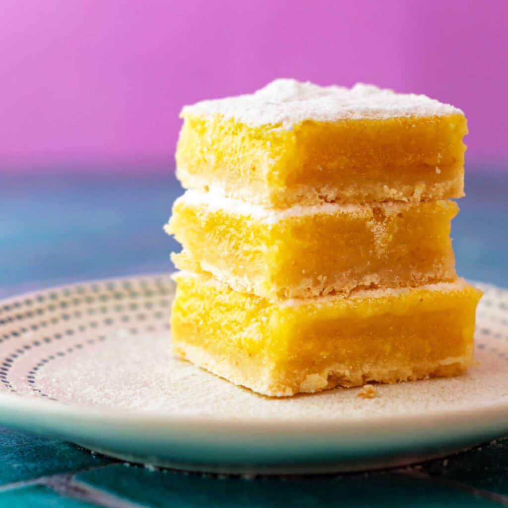 A square image of a stack of 3 square, yellow lemon bars dusted with powdered sugar on a beige plate. The background is bright pink and teal.