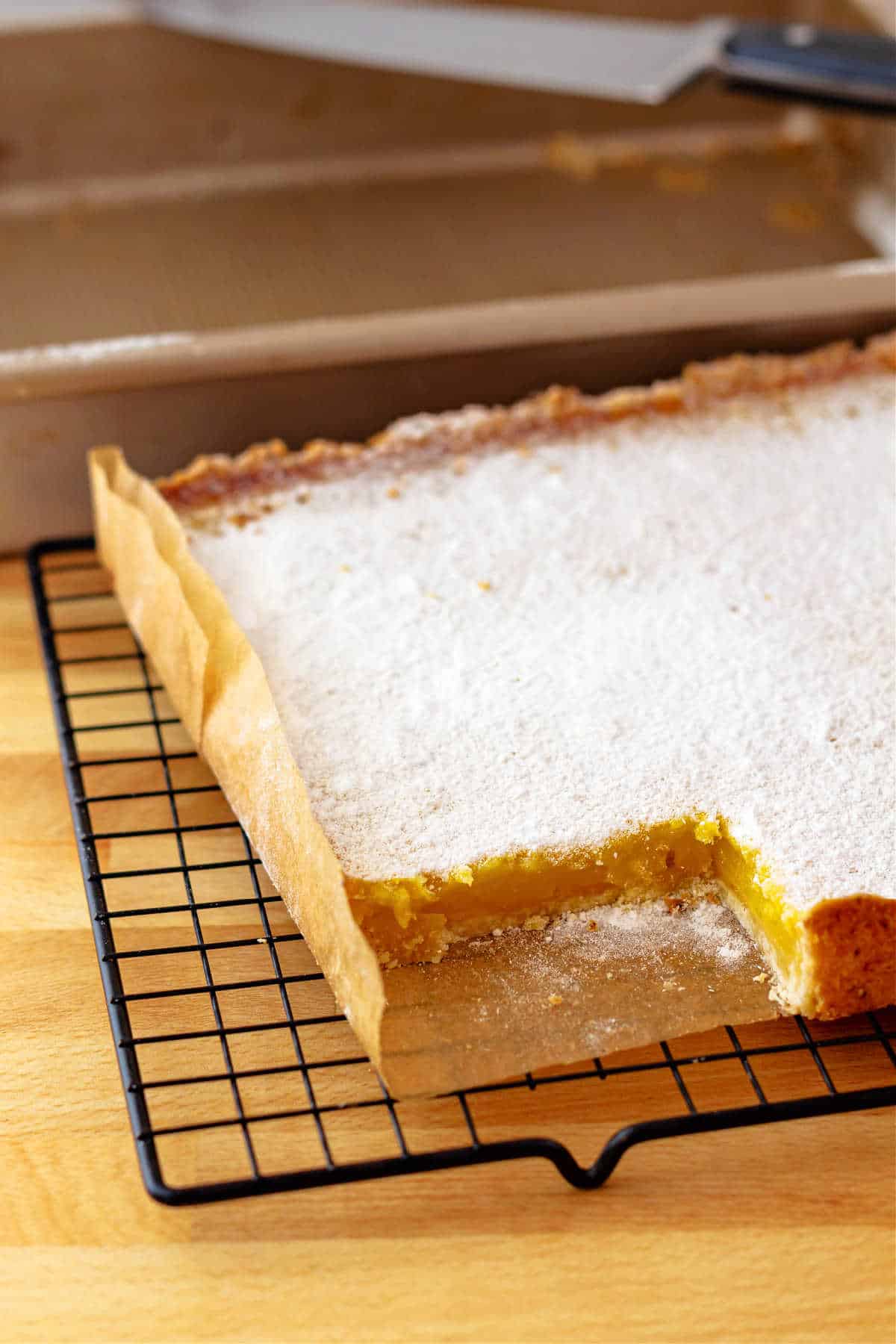 A partial view of a slab of powdered sugar-coated lemon bars with one piece cut out of it. It's on parchment on a black wire cooling rack with the baking pan in the background.