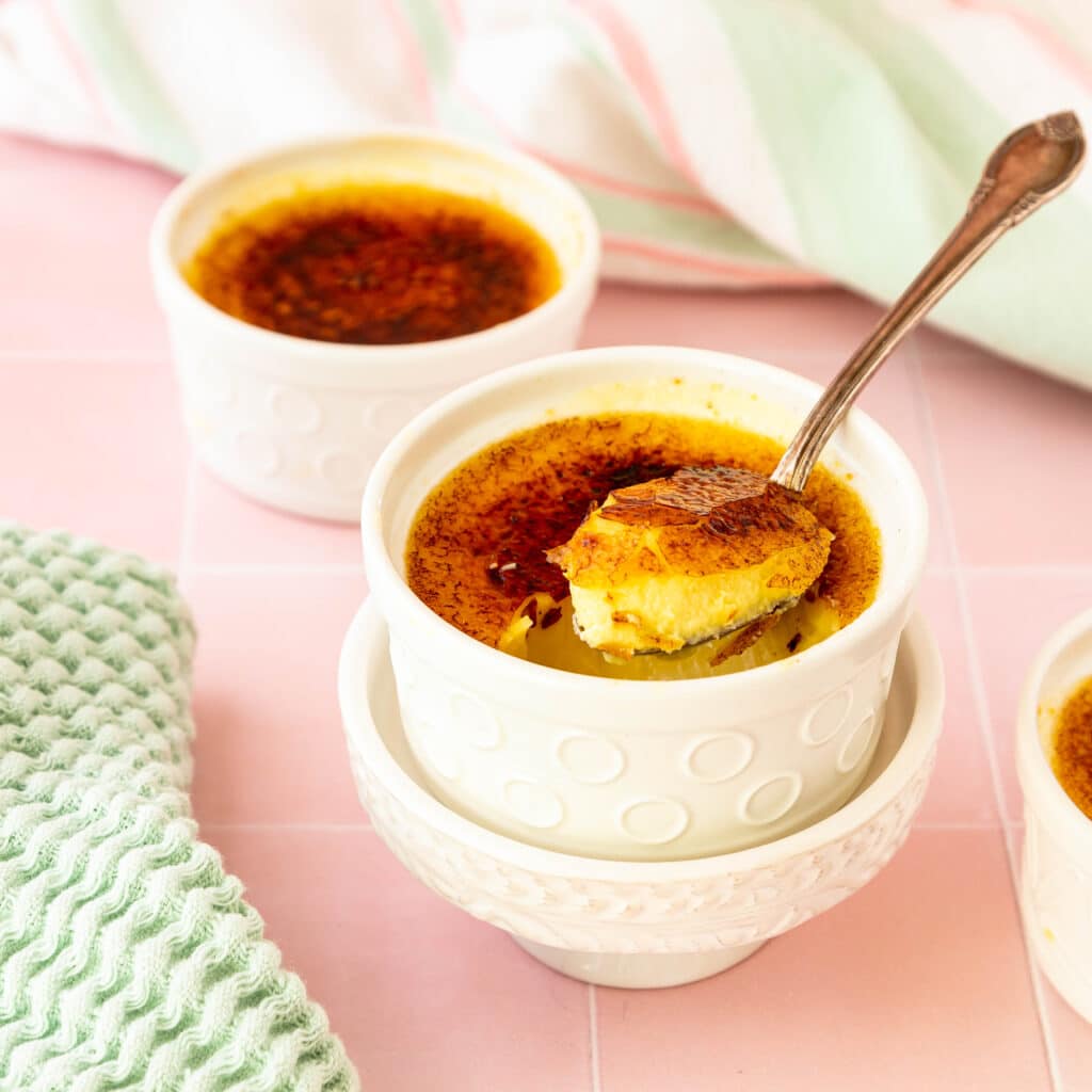Cheesecake creme brulee in a white ramekin with a spoon in it. The spoon has craqcked through the caramelized sugar shell and you can see the creamy custard underneath.