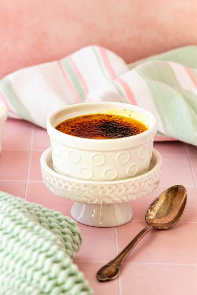 A dish of creme brulee in a white ramekin on a small white pedestal with a spoon. the background is shades of peach and pale green.